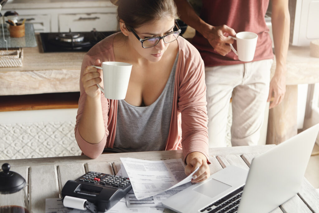 young couple calculating family expenses at home. woman in spectacles paying utility bills online, having coffee or tea, sitting in kitchen with documents and calculator, looking at laptop screen